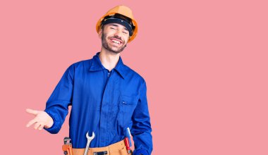 Genç İspanyol adam işçi üniforması giyiyor. Neşeli gülümsüyor ve kollarını açıyor. Hoş geldin, olumlu ve kendinden emin selamlar. 