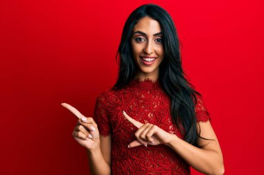 Güzel İspanyol kadın kırmızı arka planda şık kıyafetler giyiyor gülümsüyor ve kameraya bakıyor iki eliyle ve parmaklarıyla kenarı işaret ediyor. 