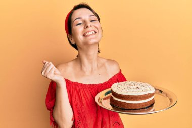 Young brunette woman with short hair holding carrot cake screaming proud, celebrating victory and success very excited with raised arm  clipart
