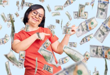 Down sendromlu esmer kadın sıradan kıyafetler giyiyor ve gözlük takıyor gülümsüyor ve kameraya bakıyor iki eliyle ve parmaklarıyla yan tarafa bakıyor..