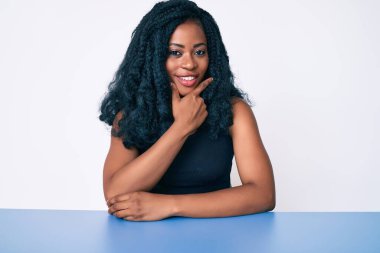 Güzel Afrikalı kadın gündelik giysiler içinde masaya oturmuş kameraya kendinden emin bir şekilde bakıyor kollarını kavuşturmuş ve çenesini kaldırmış gülümsüyor. Olumlu düşünmek. 