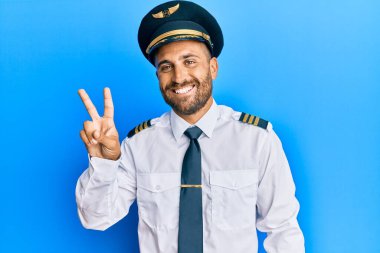 Sakallı, uçak pilotu üniforması giyen yakışıklı bir adam iki numarayı gösterip işaret ederken kendinden emin ve mutlu gülümsüyor.. 