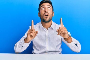 Masada oturan yakışıklı İspanyol iş kıyafetleri giymiş şaşkın ve şaşırmış bir şekilde yukarı bakıyor ve parmaklarıyla işaret ediyor ve kollarını kaldırıyordu.. 