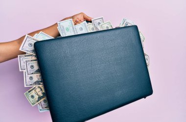 Soyutlanmış pembe arka planda dolarlarla dolu evrak çantasını tutan İspanyol adamın eli..