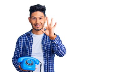 Yakışıklı Latin Amerikalı genç adam elinde antika bir telefon tutuyor parmaklarıyla imza atıyor, gülümsüyor ve mükemmel bir sembol oluşturuyor. 