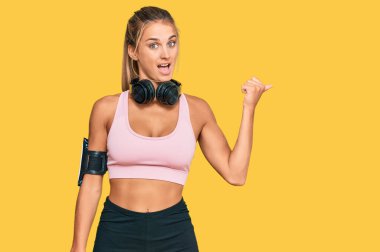 Spor kıyafetleri giyen, kulaklık takan mutlu yüzlü gülümseyen ve baş parmağıyla yüzünü gösteren genç sarışın kadın.. 