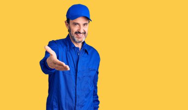 Middle age handsome man wearing mechanic uniform smiling cheerful offering palm hand giving assistance and acceptance.  clipart