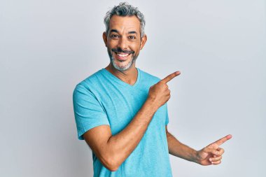 Orta yaşlı, gri saçlı, sıradan kıyafetler giyen, gülümseyen ve kameraya bakan, iki elini ve parmağını yana doğru gösteren bir adam.. 