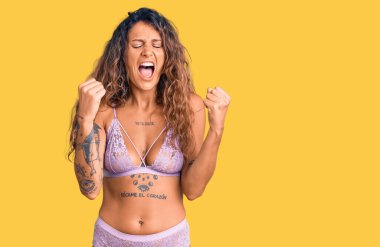 Dövmeli genç Latin bir kadın. İç çamaşırı giyiyor. Kızgın ve kızgın. Öfkeyle bağırırken yumruklarını kaldırıyor. öfke ve agresif konsept. 