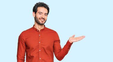 Gündelik kıyafetler giyen genç İspanyol adam neşeli bir şekilde gülümsüyor ve avuç içi kameraya bakıyor.. 