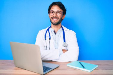 Klinikte çalışan yakışıklı, doktor üniforması giyen, kollarını kavuşturup kameraya gülümseyen yakışıklı bir adam. pozitif kişi. 