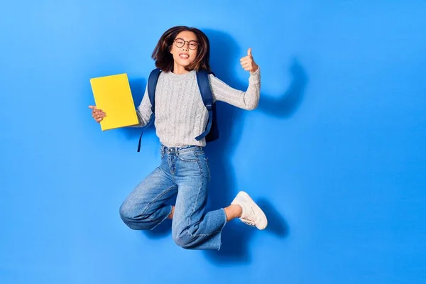 Young beautiful chinese student girl wearing glasses and backpack smiling happy. Jumping with smile on face holding book doing ok sign over isolated blue background