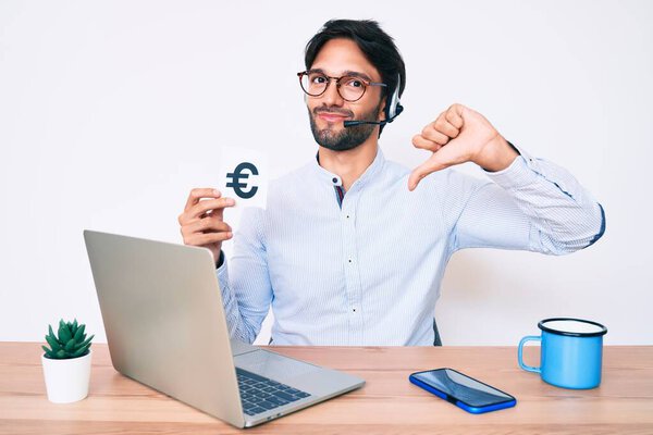 Красивый латиноамериканец, работающий в офисе, держит баннер евро с разгневанным лицом, негативный знак, показывающий неприязнь с опущенными пальцами, концепция отказа 