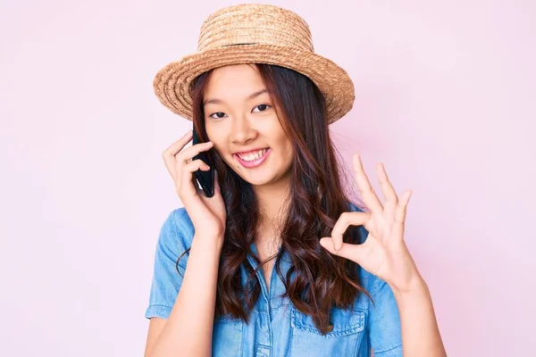 年轻美丽的中国姑娘头戴夏装 戴着夏装帽 在智能手机上聊天 用手指签着名 微笑着表示友善的手势 象征着极好的象征 — 图库照片