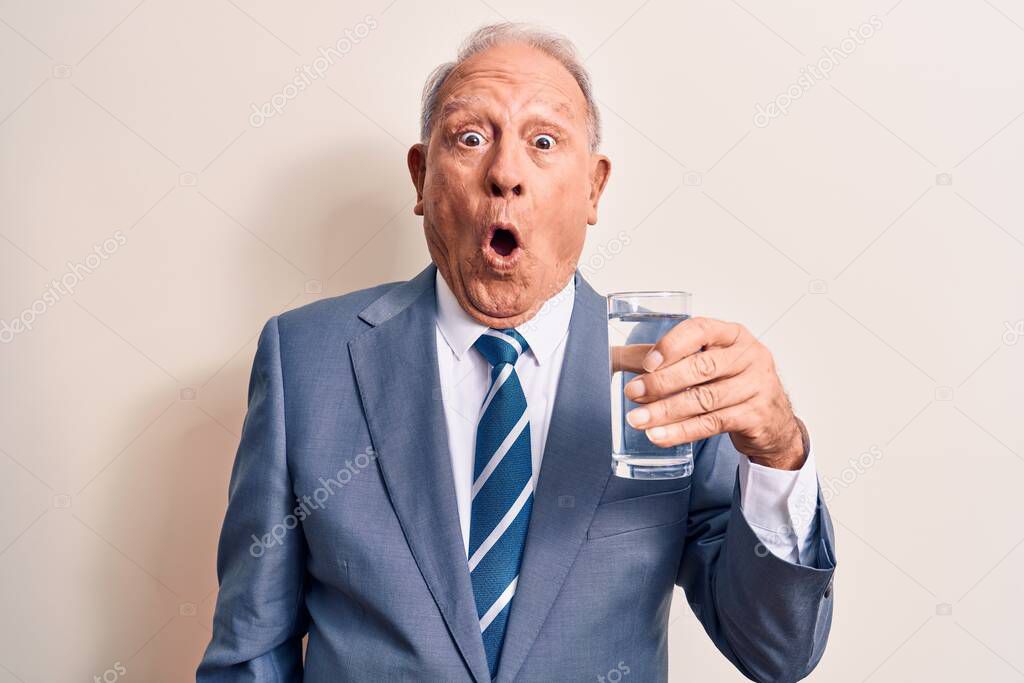 Senior handsome grey-haired businessman wearing suit drinking glass of water to refreshment scared and amazed with open mouth for surprise, disbelief face