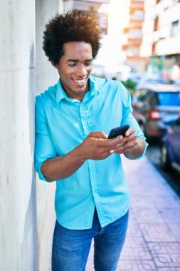 Genç, yakışıklı, Afro-Amerikan bir adam günlük giysiler giyip mutlu gülümsüyor. Yüzünde gülümsemeyle duvara yaslanıyorsun. Şehir caddesinde akıllı telefon kullanıyorsun..