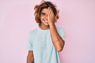 Latin asıllı genç adam gündelik giysiler giyiyor. Bir gözünü eliyle kapatıyor. Yüzünde kendine güvenen bir gülümseme ve sürpriz bir duygu var.. 