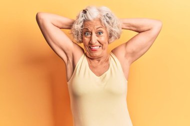 Kıdemli gri saçlı kadın rahat ve esnek giyiniyor, kolları ve elleri başının arkasında mutlu bir şekilde gülümsüyor. 