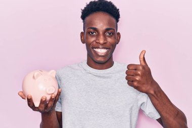 Genç Afrikalı Amerikalı domuz kumbarası tutan mutlu ve pozitif gülümseyen, başparmağıyla mükemmel ve onay işareti yapan adam. 