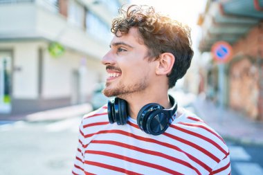 Genç beyaz adam mutlu bir şekilde gülümsüyor. Kulaklık takıyor ve şehirde yürüyor..