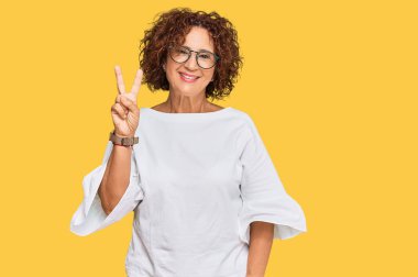 Güzel, orta yaşlı yetişkin bir kadın günlük kıyafetler ve gözlük takıyor. İki numaralı parmaklarıyla işaret ediyor ve gülümsüyor. Kendinden emin ve mutlu.. 
