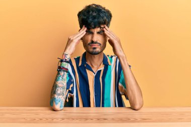 Masada oturan İspanyol asıllı genç adam acı ve migrenden dolayı çaresiz ve stresli bir şekilde baş ağrısı çekiyor. Eller başının üstünde.. 