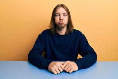 Masada oturan, uzun saçlı, günlük kıyafetler giyen, komik suratlı yakışıklı, beyaz bir adam. Havayla şişmiş ağız, çılgın bir ifade.. 