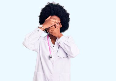 Genç Afro-Amerikalı kadın doktor önlüğü ve steteskop giyip gözlerini ve ağzını elleriyle kapatmış, şaşırmış ve şok olmuş. duygu gizleme 