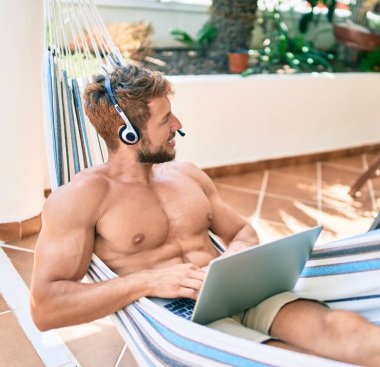 Yakışıklı beyaz adam mutlu bir şekilde gülümsüyor terasta bir hamakta dinleniyor ve çağrı merkezi kulaklığı takıyor.