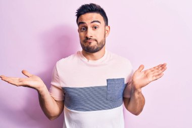 Sakallı, sıradan bir tişört giyen, pembe arka planda duran, kafası karışık, kolları açık, hiçbir fikri olmayan, kuşkulu yüzlü genç bir adam..