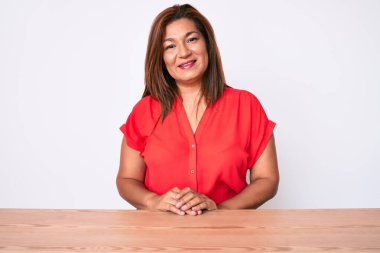 Orta yaşlı esmer, sıradan kıyafetler giyen, mutlu ve havalı bir gülümsemeyle masada oturan Latin bir kadın. şanslı kişi. 