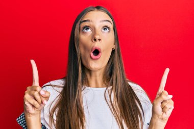 Gündelik kıyafetler giyen genç İspanyol kadın şaşırmış ve şaşırmış bir şekilde yukarı bakıyor ve parmaklarıyla işaret ediyor ve kollarını kaldırıyor.. 