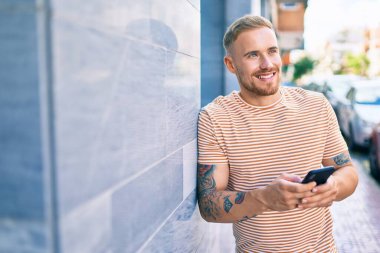 Genç İrlandalı mutlu bir şekilde gülümsüyor. Şehrin duvarına yaslanmış akıllı telefon kullanıyor..