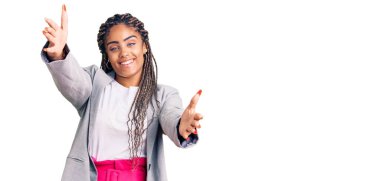 Saçları örgülü genç Afro-Amerikan kadın kameraya sarılıp gülümsüyor. Mutluluğu kucaklayan neşeli bir ifade.. 