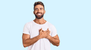 Gündelik beyaz tişört giyen, elleri göğsünde, gözleri kapalı gülümseyen ve yüzünde minnet ifadesi olan genç İspanyol bir adam. sağlık kavramı. 