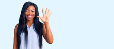 Genç Afro-Amerikalı kadın günlük giysiler giyip beş numaralı parmaklarıyla işaret ederken kendinden emin ve mutlu bir şekilde gülümsüyor.. 