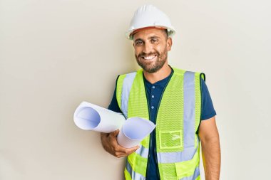 Güvenlik kaskı takan sakallı yakışıklı adam elinde planlar var. Olumlu ve mutlu görünüyor. Kendine güvenen bir gülümsemeyle dişlerini gösteriyor. 