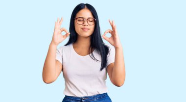 Genç ve güzel Asyalı kız günlük kıyafetler ve gözlük takıyor rahatlamış ve gözleri kapalı gülümsüyor parmaklarıyla meditasyon hareketi yapıyor. yoga konsepti. 
