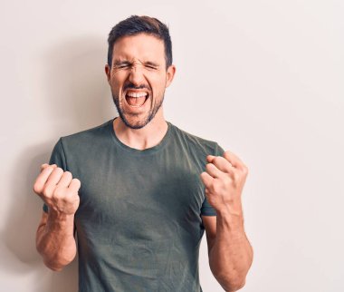 Genç ve yakışıklı bir adam, günlük tişört giyip izole edilmiş beyaz arka planda dikiliyor. Başarının şaşkınlığını ve şaşkınlığını kutluyor. Kolları havada, gözleri kapalı.