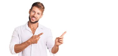 Gündelik giysiler giymiş genç beyaz bir adam gülümseyip kameraya bakıyor ve iki eliyle ve parmaklarıyla kenarı işaret ediyor.. 