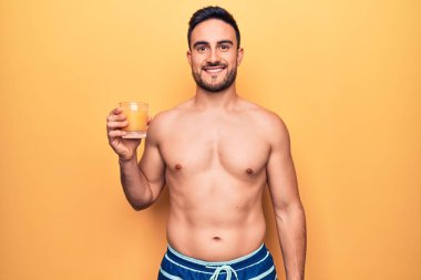 Tatilde sakallı, mayo giyen, bir bardak portakal suyu içen, pozitif ve mutlu görünen, kendine güvenen, gülümseyen, dişlerini gösteren genç bir adam.