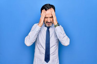Orta yaşlı, yakışıklı iş adamı zarif bir kravat takıyor. İzole edilmiş mavi arka plan üzerinde duruyor. Elleri başının üstünde, stres yüzünden başı ağrıyor. Migren ağrısı..