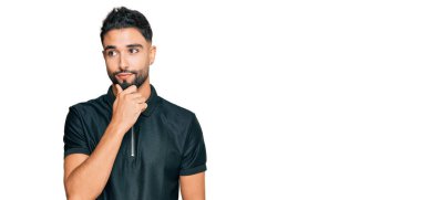 Sakallı, spor giyimli, çenesi çenesinde soru soran, düşünceli bir genç. Düşünceli bir yüzle gülümsüyor. şüphe kavramı. 