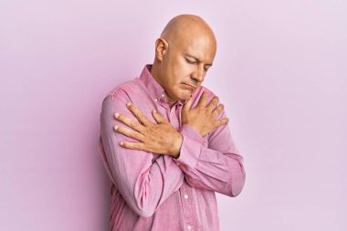 Orta yaşlı kel bir adam pembe iş gömleği giyip kendine sarılıyor mutlu ve pozitif, kendinden emin gülümsüyor. Sevgi ve özgüven. 