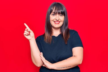 Genç ve cüsseli kadın gündelik kıyafetler giyiyor. Gülümsüyor, mutlu bir şekilde elini ve parmağını yana doğru gösteriyor. 