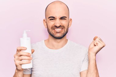 Genç yakışıklı kel adam elinde krem losyon şişesiyle gururla bağırıyor, zaferi kutluyor ve başarıyı kollarını kaldırarak kutluyor. 