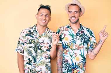 Genç eşcinsel iki adam yaz şapkası takıyor ve Hawaii gömleği giyiyor. Yüzünde kocaman bir gülümseme var. Elleriyle kameraya bakıyor.. 