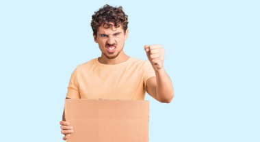 Genç, kıvırcık saçlı, elinde boş karton pankart olan yakışıklı bir adam. Sinirli ve sinirli bir şekilde bağırıyor. Öfkeyle bağırıyor ve el kaldırıyor. 