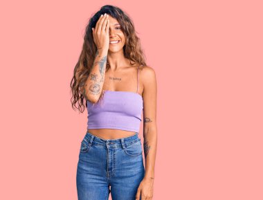 Dövmeli genç İspanyol bir kadın, günlük kıyafetler giyiyor, bir gözünü eliyle kapatıyor, yüzünde kendine güvenen bir gülümseme ve sürpriz bir duygu var.. 