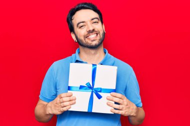 Genç İspanyol adam elinde hediye tutuyor. Pozitif ve mutlu görünüyor. Kendine güvenen bir gülümsemeyle ayakta duruyor ve dişlerini gösteriyor. 